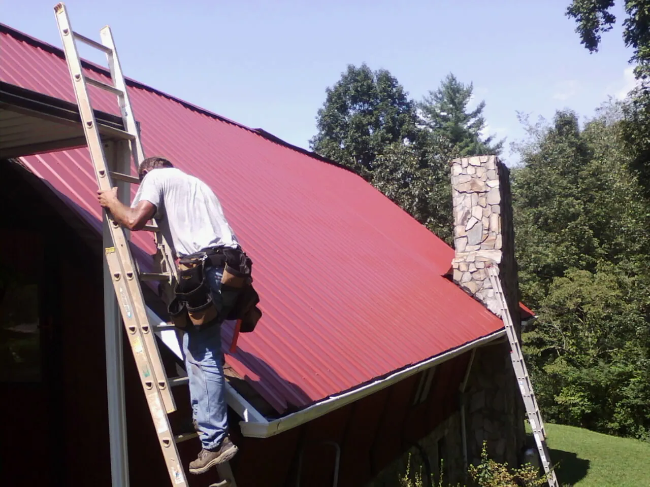 Roofer on a ladder beside a red metal roof and stone chimney — Soffit & Fascia Repair by Roofing Brothers in Galion